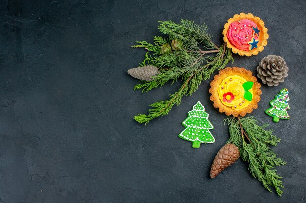 Вид сверху маленькие пирожные, рождественское печенье, сосновые шишки, сосновые ветки на темном столе с копией пространства