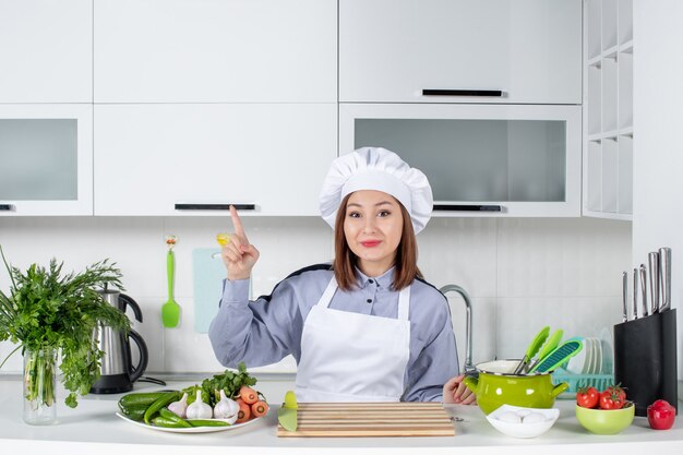 Top view of smiling concentrated female chef and fresh vegetables pointing up in the white kitchen