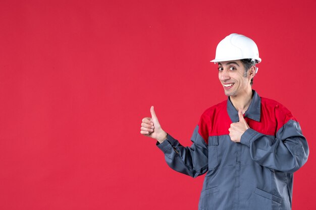 모자를 쓴 제복을 입고 웃고 있는 자신감 있는 젊은 노동자의 상위 뷰와 격리된 붉은 벽에 확인 제스처를 하는 모습
