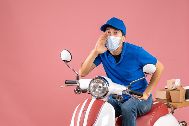 Top view of smiling courier man in medical mask wearing hat sitting on scooter calling someone on pastel peach