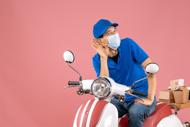 Top view of smiling courier man in medical mask wearing hat sitting on scooter listening to the last gossiping on pastel peach