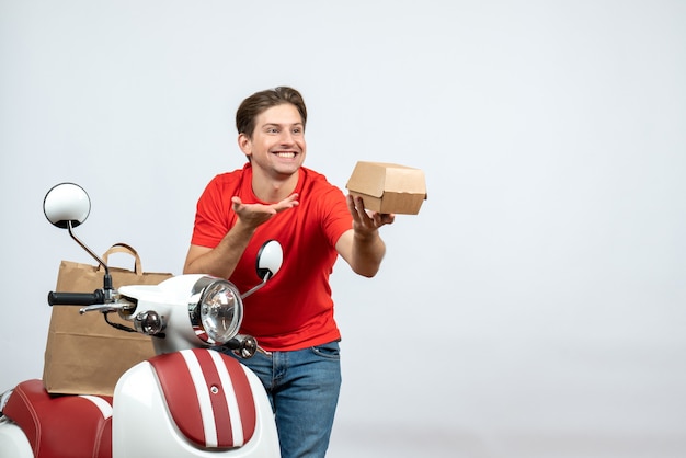 Top view of smiling delivery guy in red uniform standing near scooter holding order and looking at something on white wall