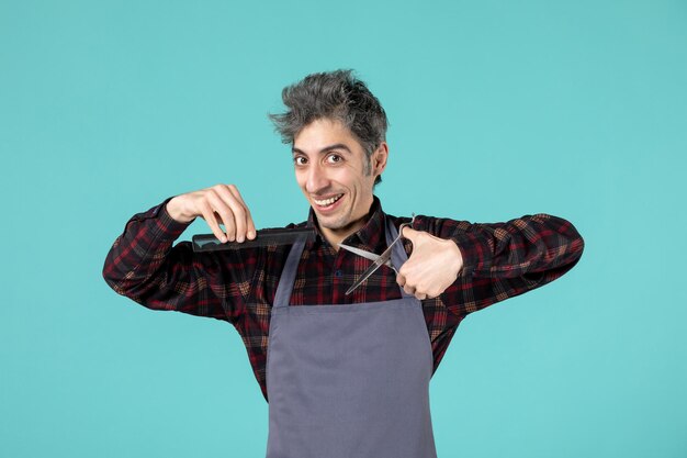 Top view of smiling emotional young hairdesser wearing gray apron and holding comb and scissor on pastel blue color surface