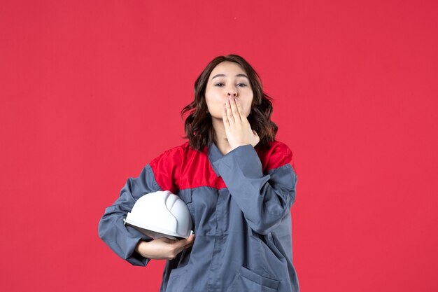 Top view of smiling female builder in uniform and holding hard hat making kiss gesture on isolated red background