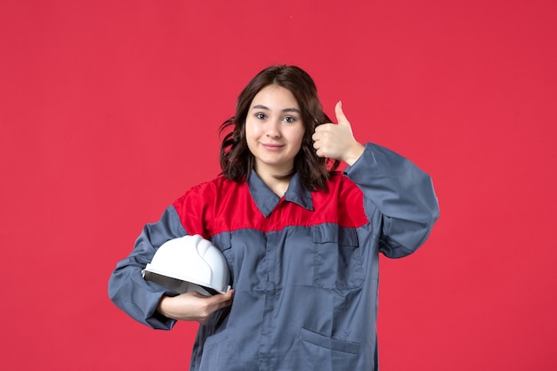Top view of smiling female builder in uniform and holding hard hat making ok gesture on isolated red background