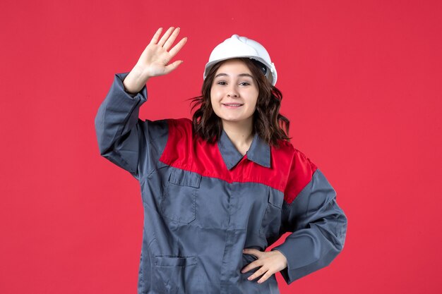 Top view of smiling female builder in uniform with hard hat saying hello to someone on isolated red background