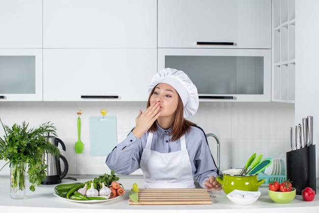 Vista dall'alto della chef donna sorridente e verdure fresche con attrezzature da cucina e che fa il gesto del bacio a qualcuno nella cucina bianca