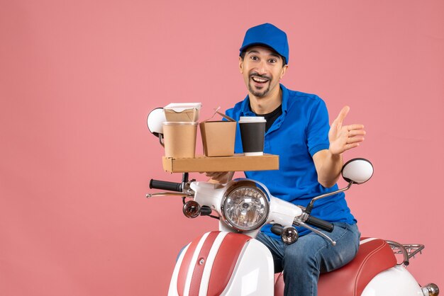 Top view of smiling happy courier man wearing hat sitting on scooter on pastel peach