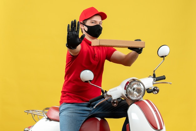 Top view of smiling happy courier man wearing red blouse and hat gloves in medical mask sitting on scooter showing order showing five