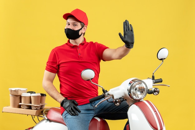 Top view of smiling happy young adult wearing red blouse and hat gloves in medical mask delivering order sitting on scooter showing five on yellow wall