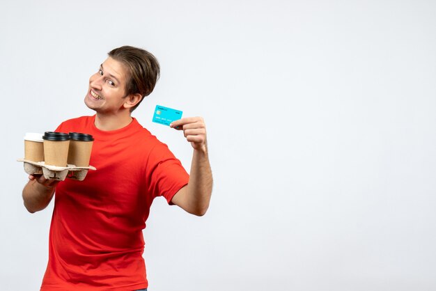 Top view of smiling and happy young man in red blouse holding orders bank card on white wall