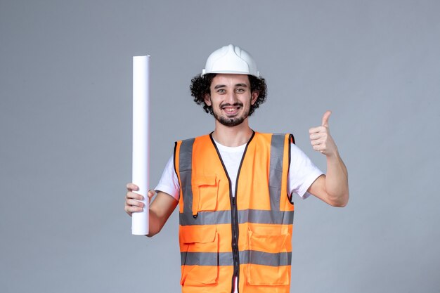 Top view of smiling male constructor in warning vest wearing safety helmet and holding blank making ok gesture on gray wave wall