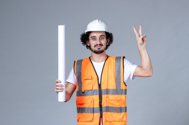 Top view of smiling male constructor in warning vest wearing safety helmet and holding blank making victory gesture on gray wave wall