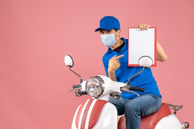 Top view of smiling male delivery person in mask wearing hat sitting on scooter showing document on pastel peach