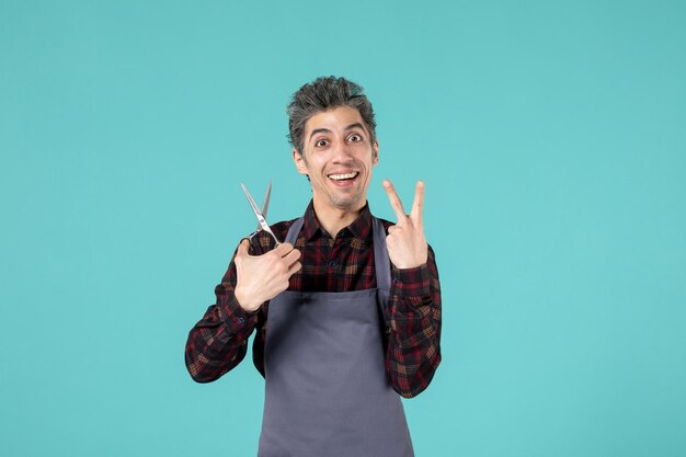 Top view of smiling male hairdesser wearing gray apron and holding scissor making victory gesture on blue soft color background