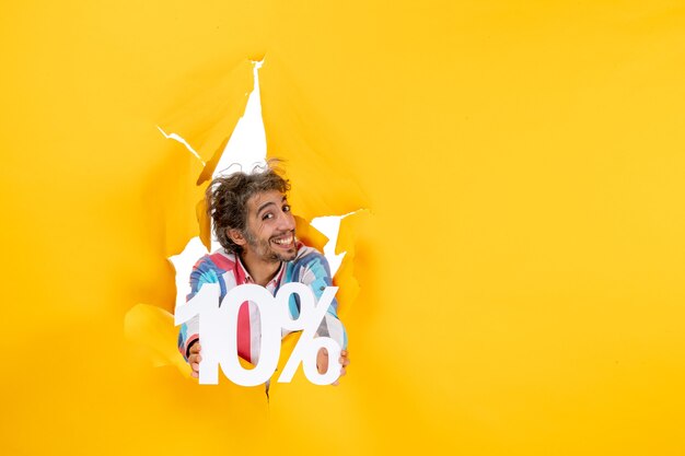 Top view of smiling and positive young man showing ten percentage in a torn hole in yellow paper