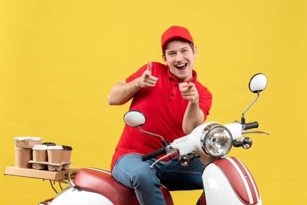 Top view of smiling young adult wearing red blouse and hat delivering order sitting on scooter pointing forward on yellow wall