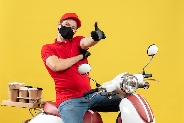 Vista superiore del giovane adulto sorridente che indossa la camicetta rossa e guanti del cappello nella mascherina medica che trasporta l'ordine che si siede sullo scooter che fa il gesto giusto sulla parete gialla