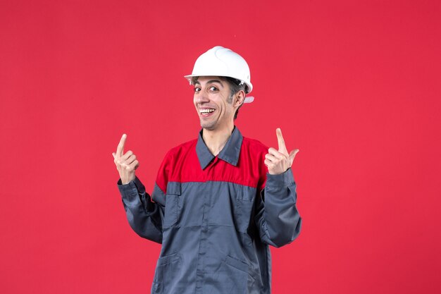 Top view of smiling young builder in uniform wearing hard hat pointing up on isolated red wall