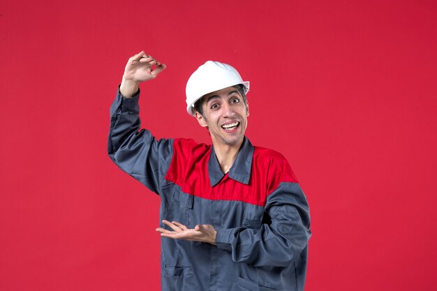 Top view of smiling young builder in uniform with hard hat explaining something on isolated red wall