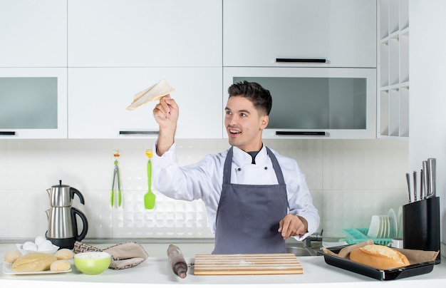 Top view of smiling young cook in uniform standing behind the table in the white kitchen