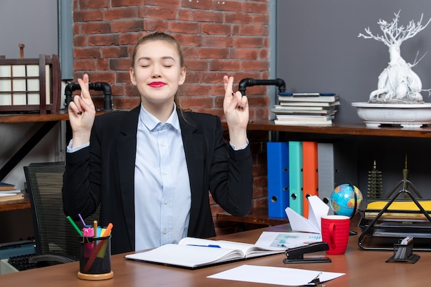 Top view of smiling young female sitting at a table and holding the document crossing her fingers dreaming about something in the office