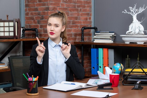 Top view of smiling young female sitting at a table and pointing up in the office