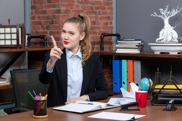 Top view of smiling young female sitting at a table and showing one in the office