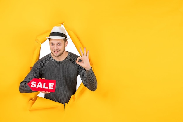 Foto gratuita vista dall'alto di un giovane ragazzo sorridente che tiene in mano un cartello di vendita e fa un gesto con gli occhiali in una parete gialla strappata yellow