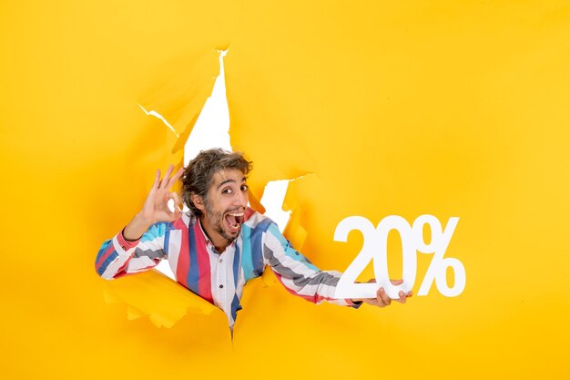 Top view of smiling young man holding twenty percentage and making eyeglasses gesture in a torn hole in yellow paper