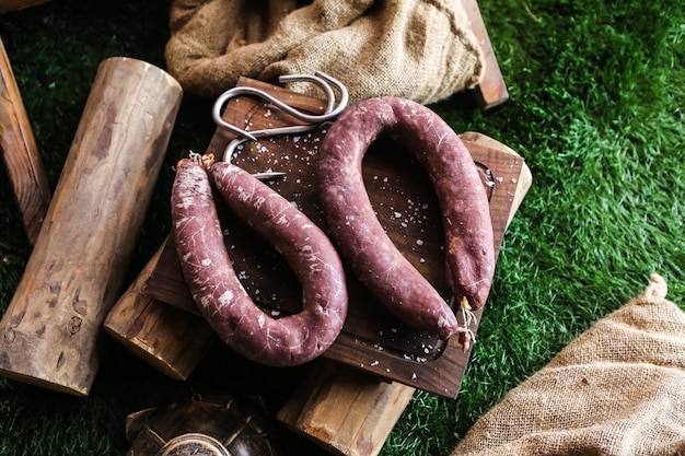 Вид сверху копченая колбаса на подносе с дровами и мешковины на траве