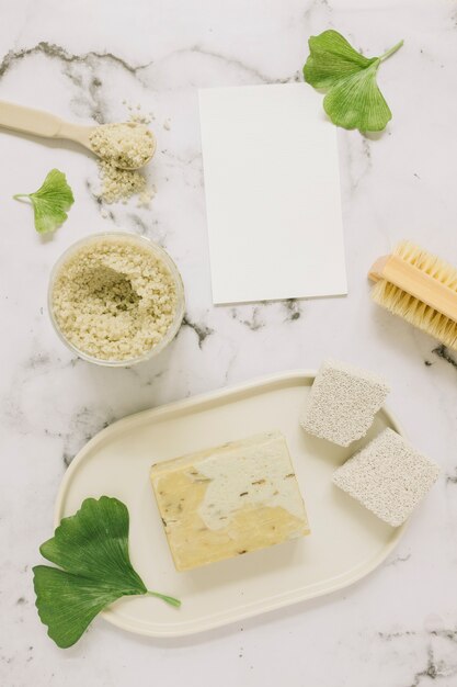 Top view of soap; salt; pumice stone; brush; ginkgo leaf and blank card on marble backdrop