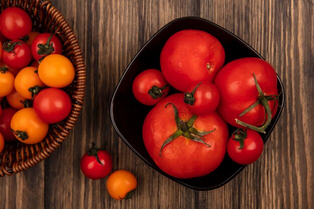 Top view of soft red and orange cherry tomatoes on a bucket with large size tomatoes on a bowl on a wooden wall