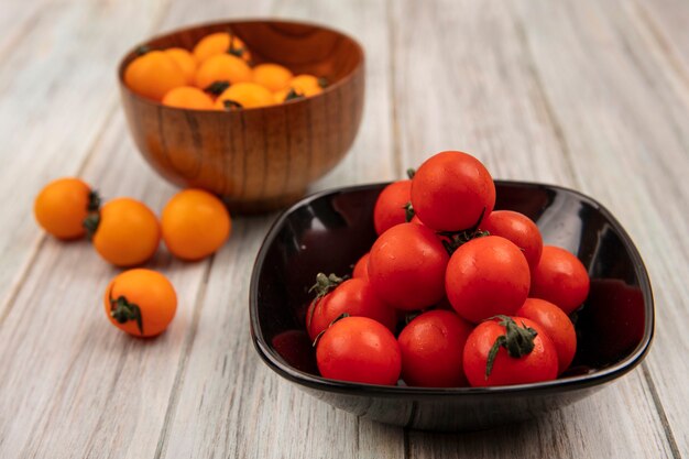 Top view of soft red tomatoes on a black bowl with orange tomatoes on a wooden bowl on a grey wooden surface