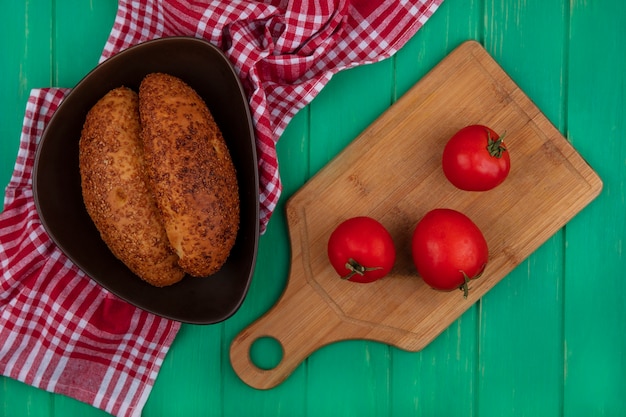 Top view of soft and sesame patties on a bowl with fresh tomatoes on a wooden kitchen board on a red checked cloth on a green wooden background