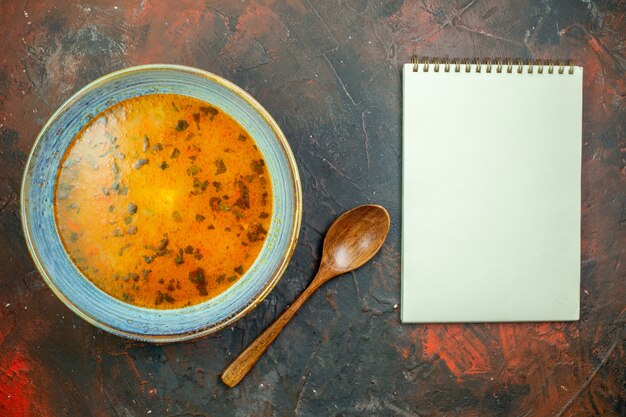 Top view soup in blue bowl wooden spoon notebook on dark red table