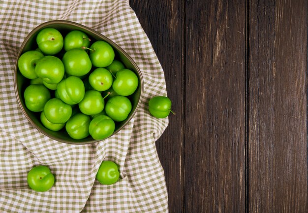 Top view of sour green plums in a bowl on plaid fabric on dark wooden table with copy space