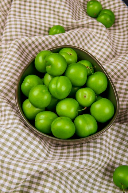 Top view of sour green plums in a bowl on plaid fabric