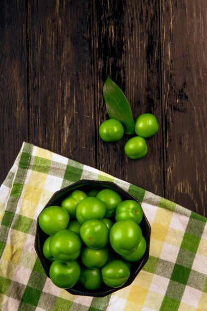 Top view of sour green plums in a bowl on plaid napkin on dark wooden table