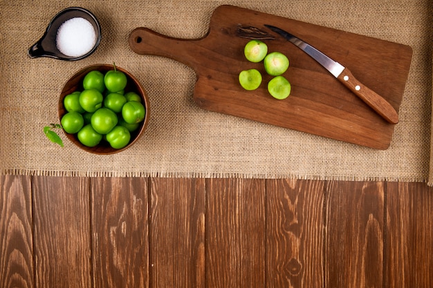 ボウルにサワーグリーンプラムとナイフで木製のまな板にスライスしたプラムとコピースペースを持つ暗い素朴なテーブルに荒布を着た塩のトップビュー