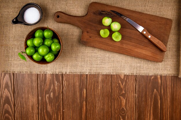 Top view of sour green plums in a bowl and sliced plums on a wooden cutting board with knife and salt on sackcloth on dark rustic table with copy space