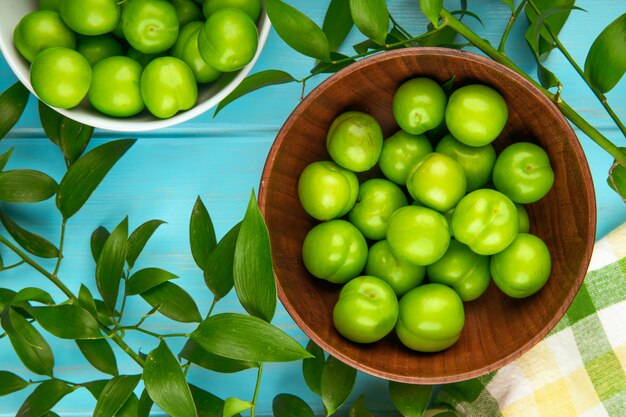 Top view of sour green plums in bowls on blue wooden table