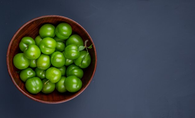 Top view of sour green plums in a wooden bowl on black table with copy space