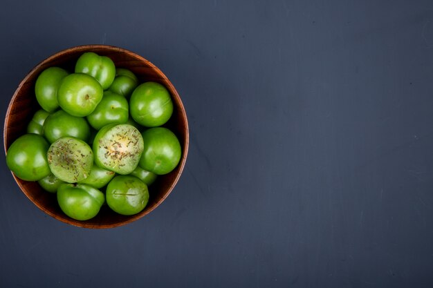 Top view of sour green plums in a wooden bowl on black table with copy space
