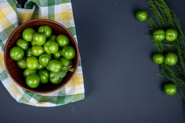 Top view of sour green plums in a wooden bowl on plaid napkin and scattered green plums with fennel on black table with copy space