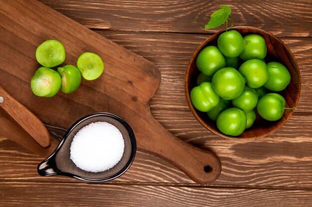 Top view of sour green plums in a wooden bowl and sliced plums with kitchen knife and salt in a saucer on rustic table