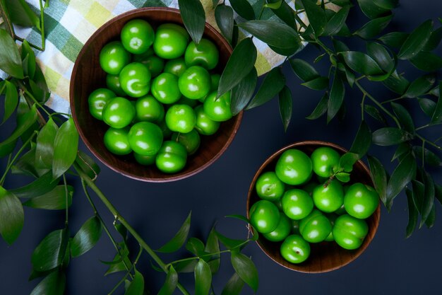 Top view of sour green plums in wooden bowls and ruscus leaves on black table