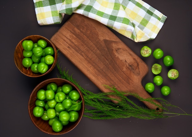 Foto gratuita vista superiore delle prugne verdi acide in ciotole di legno e un tagliere di legno con il tovagliolo del plaid sulla tavola nera