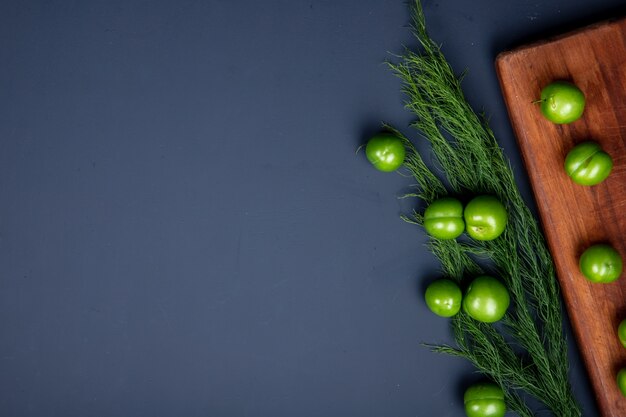 Top view of sour green plums on a wooden cutting board and fennel on black table with copy space