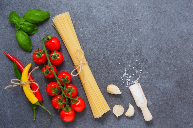 Top view spaghetti pasta with chili peppers, a bunch of tomatoes, salt, black pepper, garlic, leaves on gray background. space for text